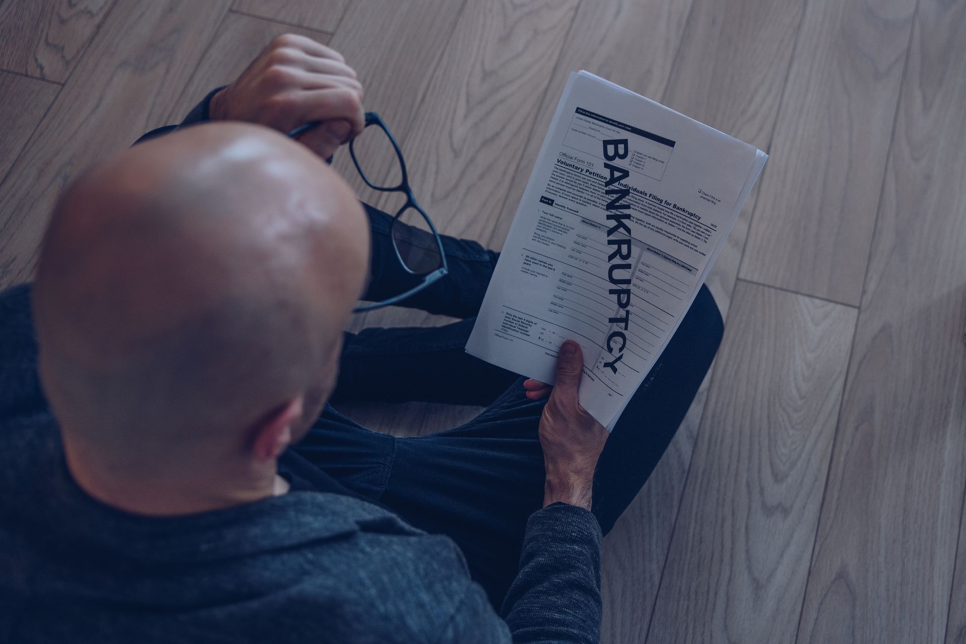 A person holds bankruptcy document, sitting on wooden floor, looking stressed.