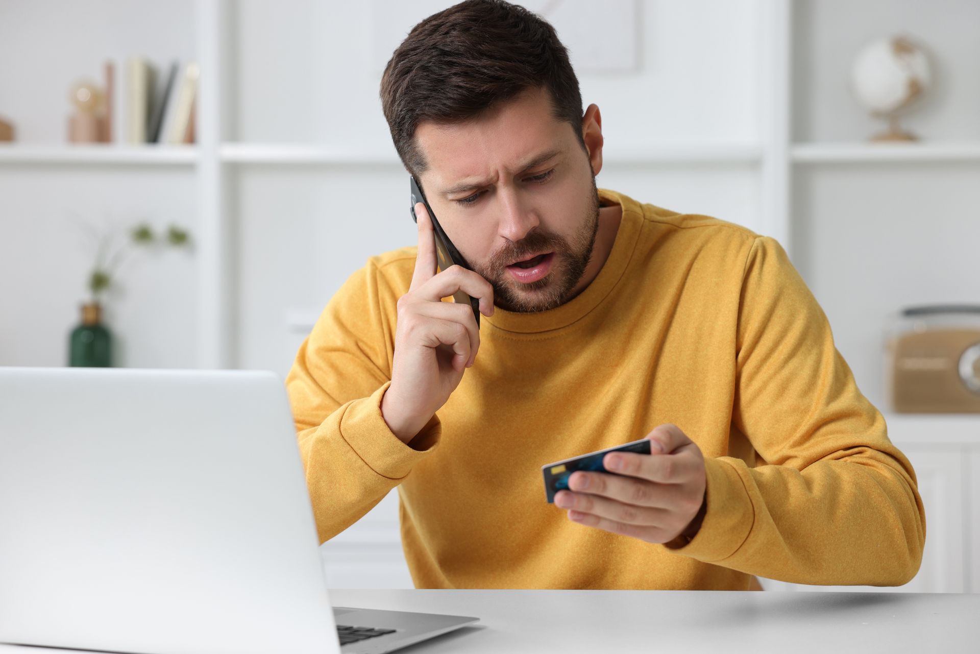 Man on phone looks concerned, holding small object, near laptop in a home office.