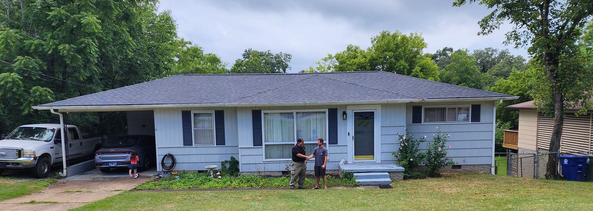A single-story, light-blue house with a carport, two people standing in the front yard, and a pickup truck parked outside.