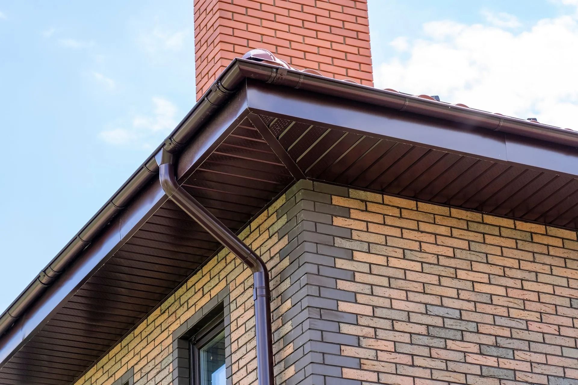 Brick house corner with brown gutters, white trim, and a red chimney against a blue sky