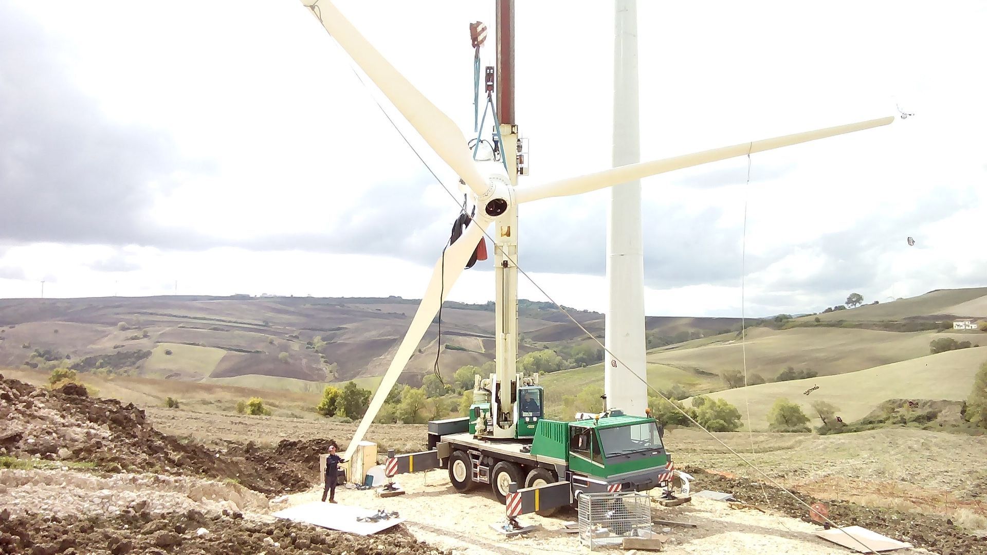 Una gru sta montando una turbina eolica bianca su una fondazione di cemento in un paesaggio rurale collinare sotto un cielo nuvoloso.