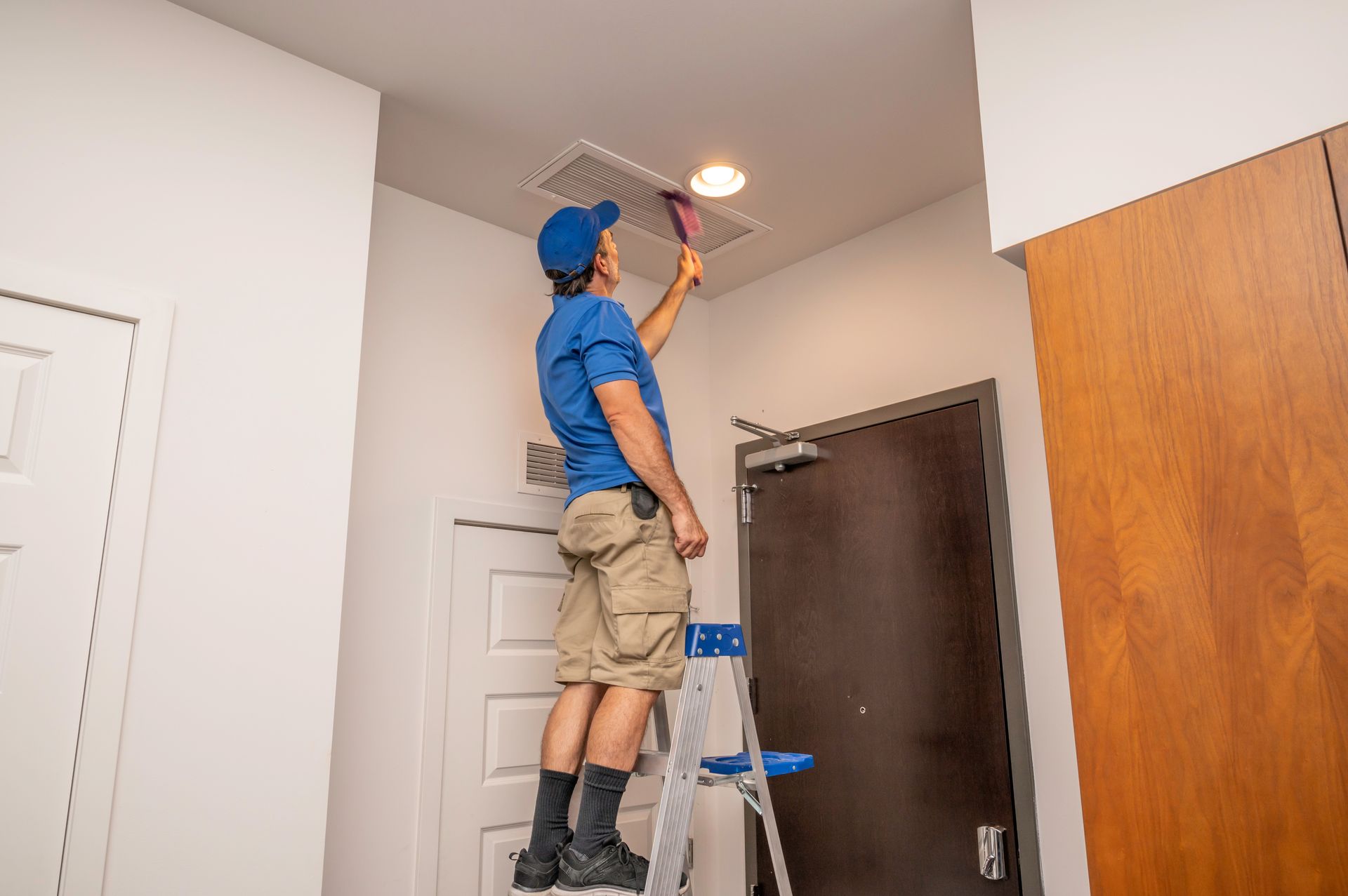 A man is standing on a ladder in a room looking at an air vent.