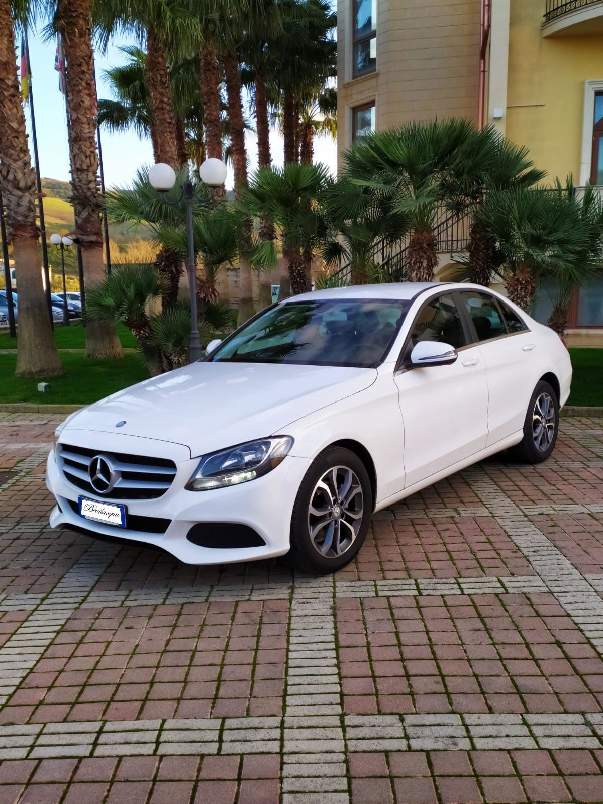 Una mercedes benz bianca è parcheggiata su un marciapiede di mattoni di fronte a un edificio.