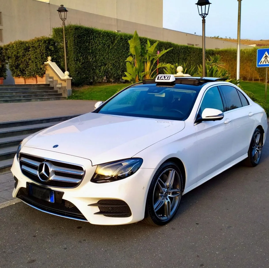 Una berlina Mercedes bianca parcheggiata davanti a un edificio con siepi e lampioni al tramonto.