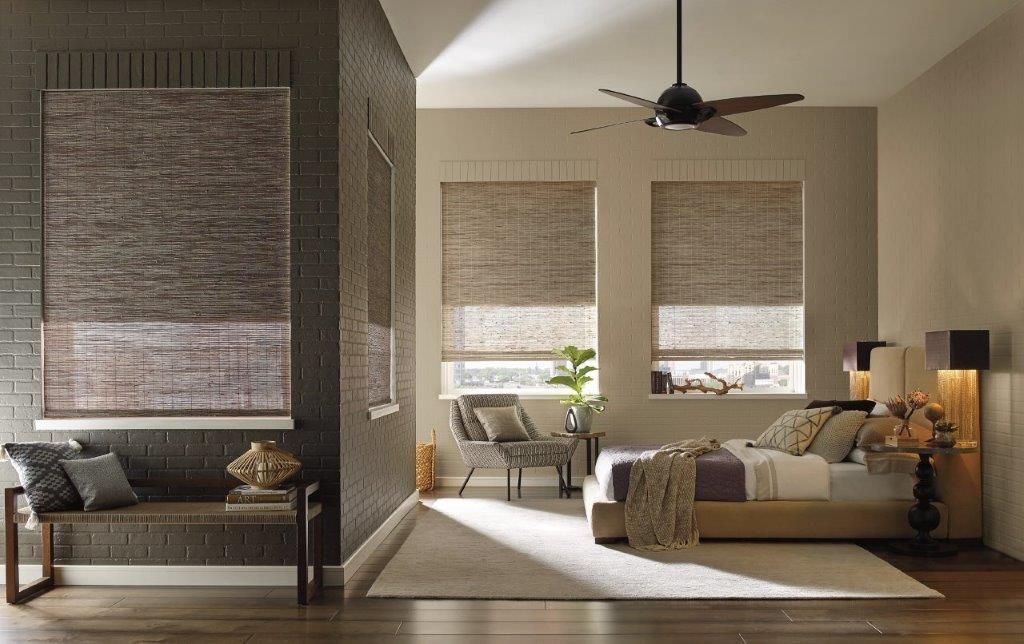 Bedroom with woven shades, brick accent wall, bench, bed, and seating area, natural light.