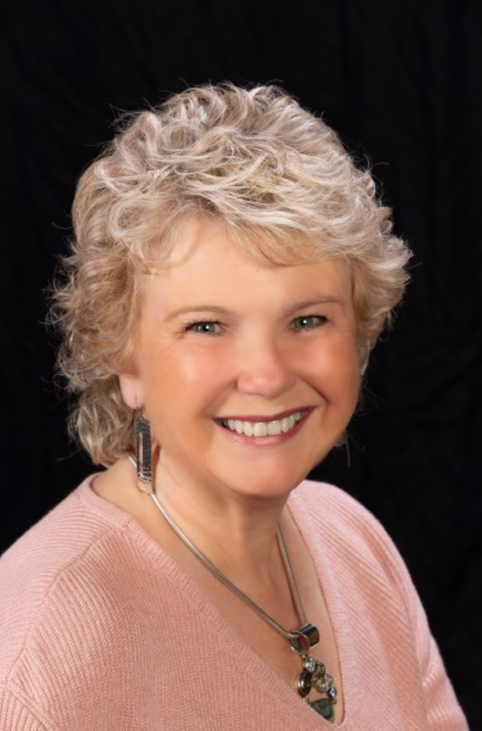 Smiling Aileen Smart with short, curly blonde hair wearing a pink sweater and jewelry. Black background.