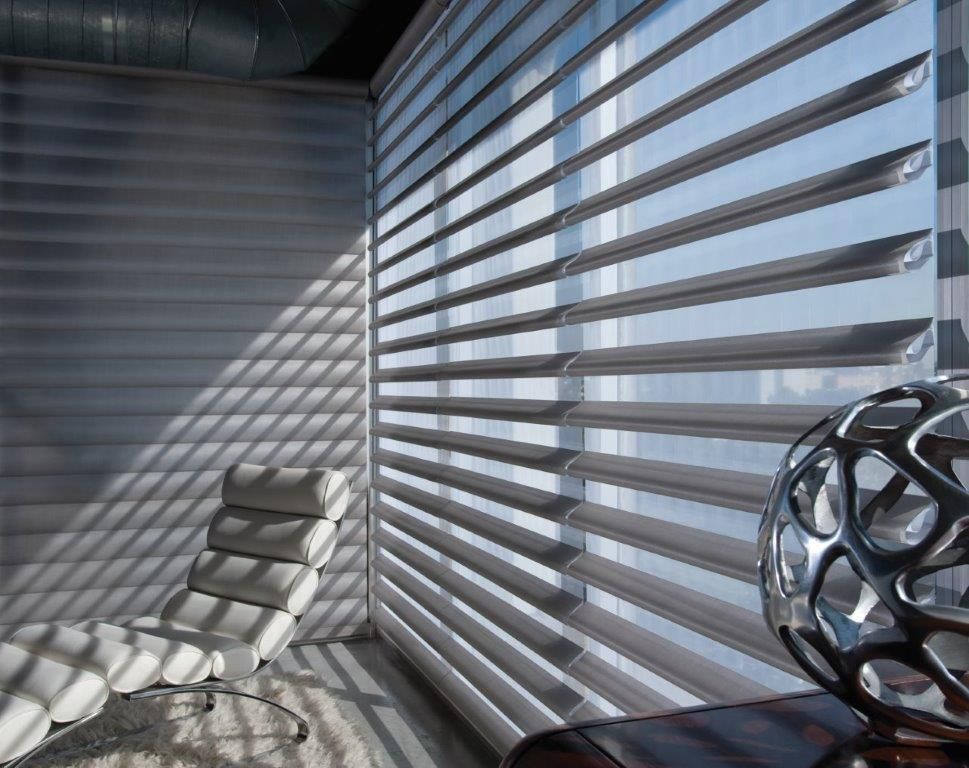 Room with a window featuring layered blinds casting shadows on a white chair and metallic sculpture.