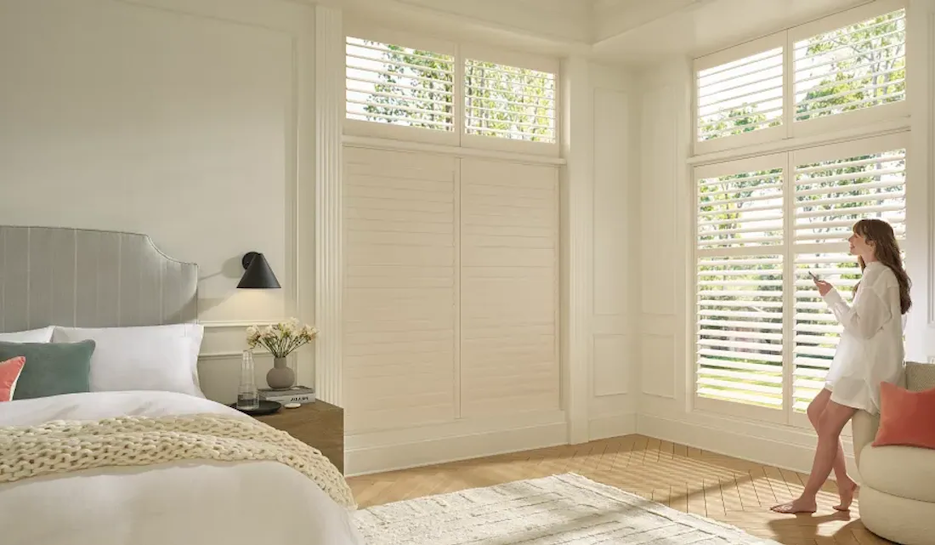 Woman adjusting white shutters in a bright bedroom with a bed and sunny windows.