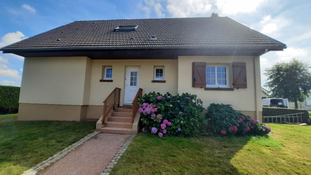 Maison beige avec volets marron, porte d'entrée et marches marron, et fleurs roses dans le jardin.