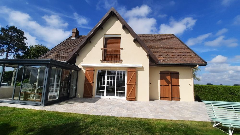 Maison beige avec volets et toit marron, véranda et ciel bleu.