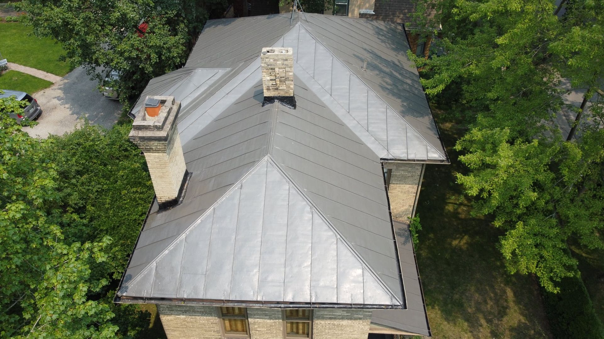 Overhead view of a house with a gray metal roof, chimneys, and surrounding green trees.