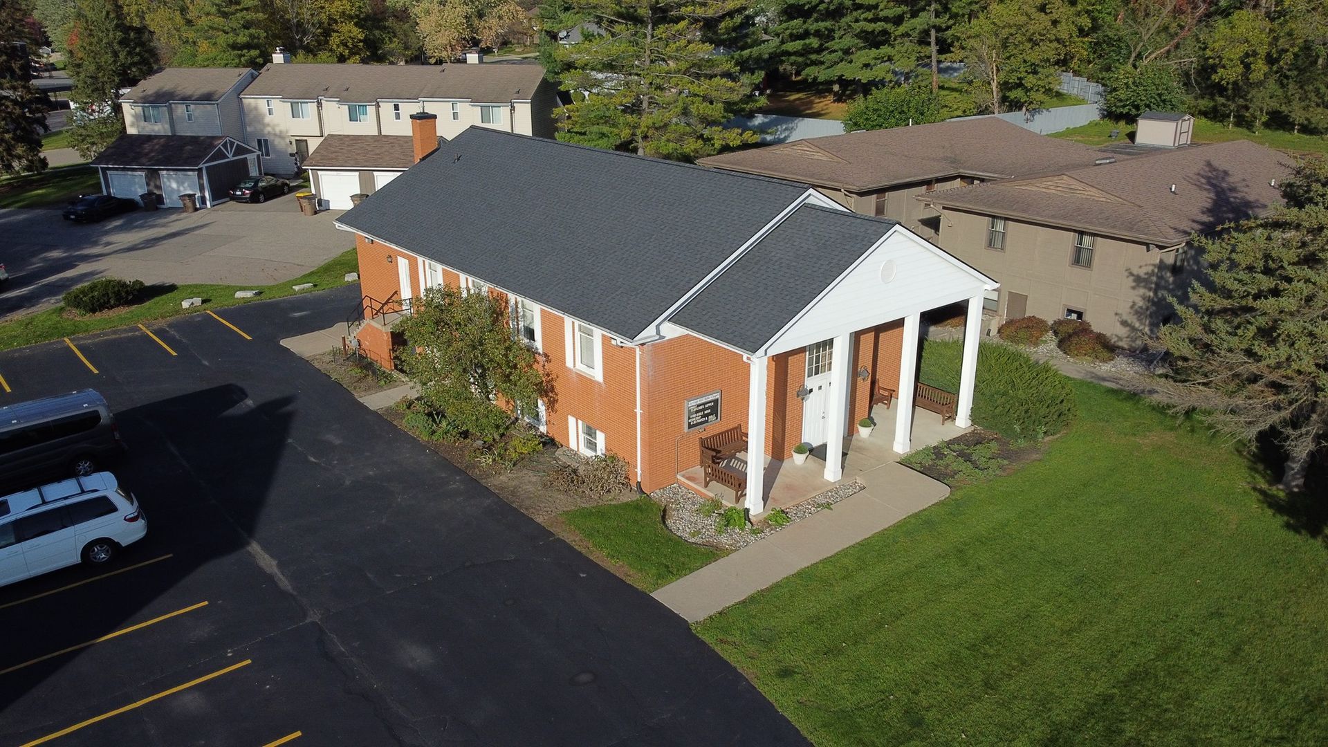 Brick building with a dark gray roof and white columns, surrounded by grass and a parking lot.