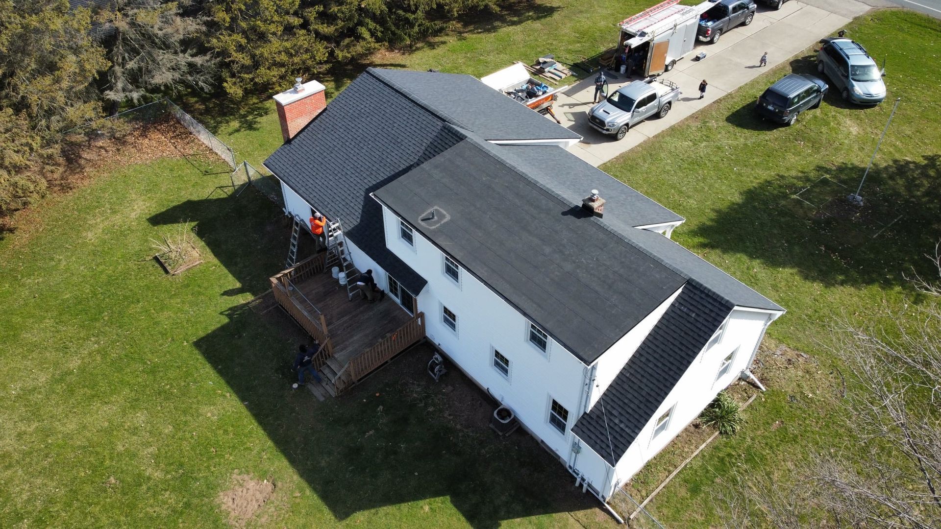 Aerial view of a white two-story house with a black roof. Construction crew on the roof, with vehicles nearby on green grass.