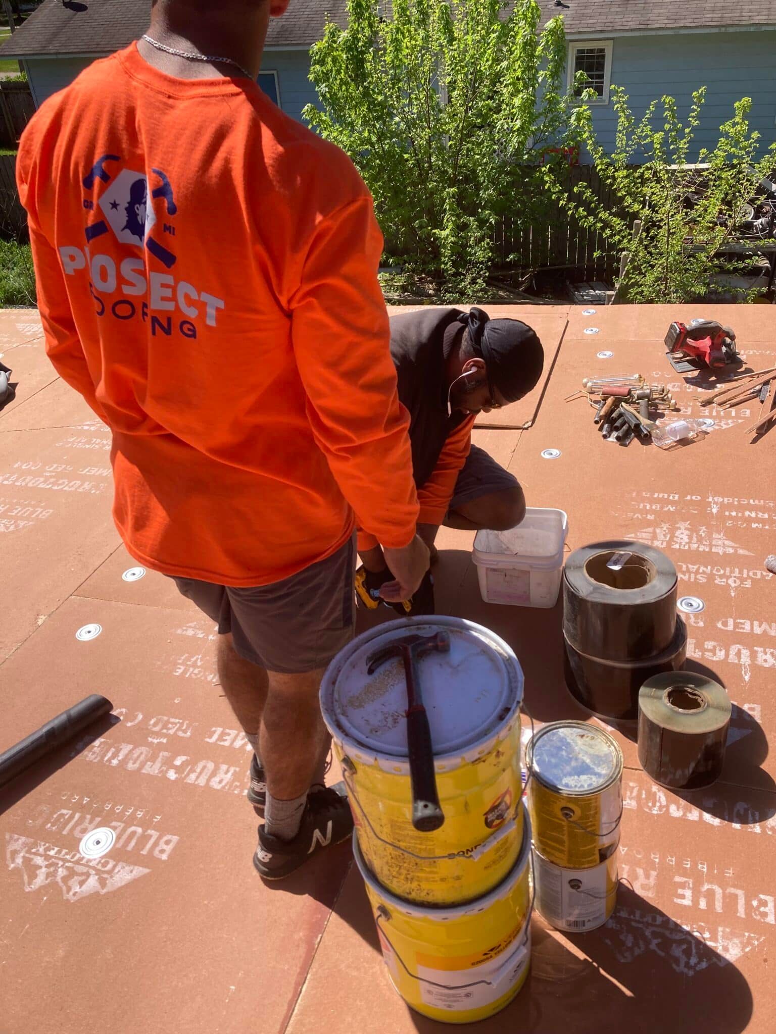 Two people working on a roof; one holds tools, others assemble material. Orange shirt, yellow buckets, setting sun.
