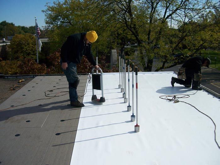 Two workers on a rooftop installing white roofing material, one holding a device.