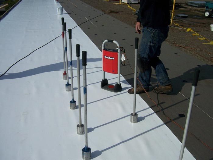 Person using tools to test a white membrane roofing material, near a red cart on a rooftop.