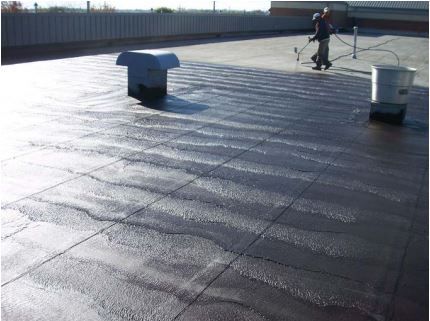 Person spraying a dark substance on a flat roof. A vent and bucket are also on the roof.