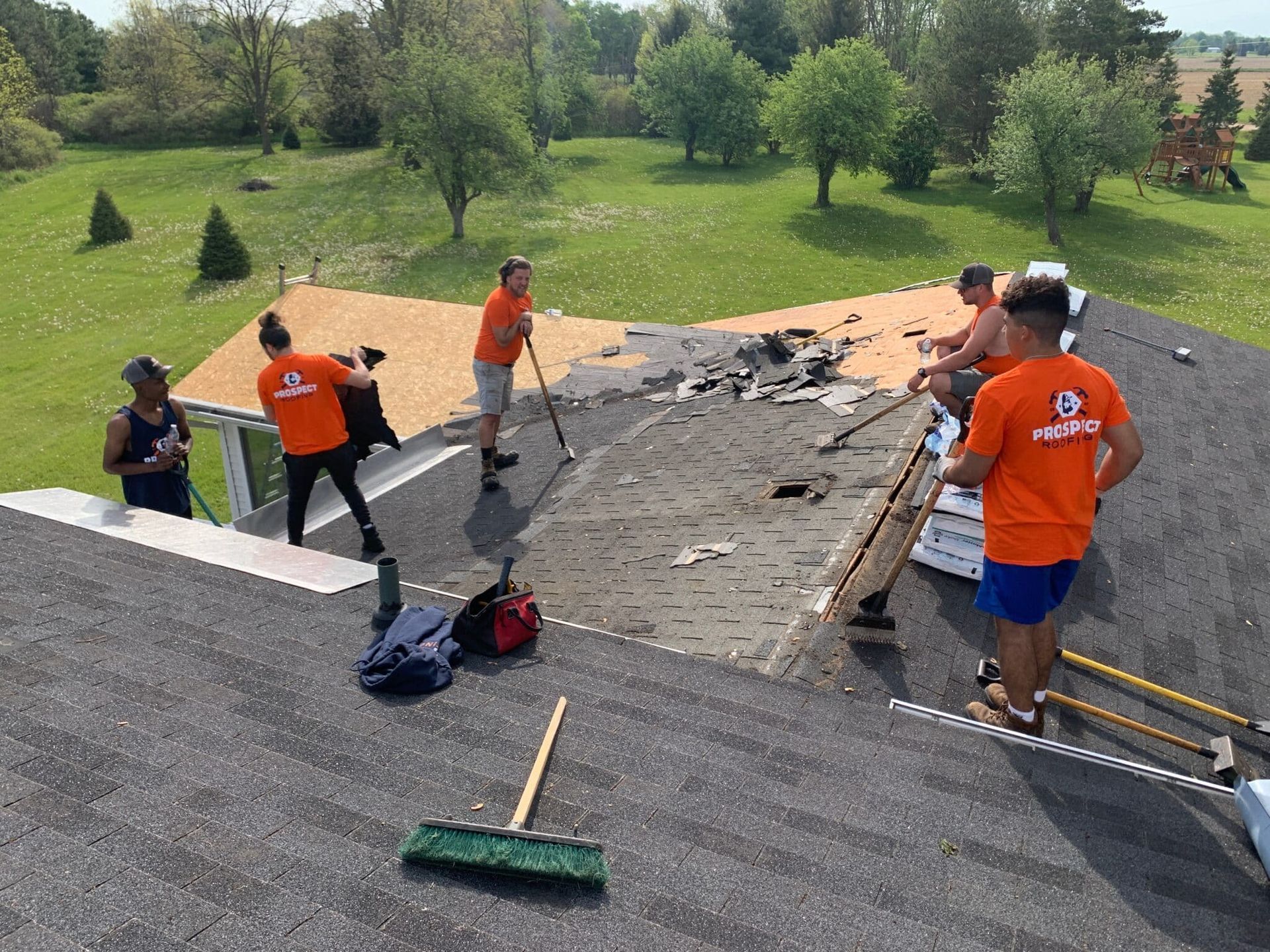 Roofing crew removing shingles on a sunny day. Men in orange shirts work with tools on the roof. Green grass and trees in background.