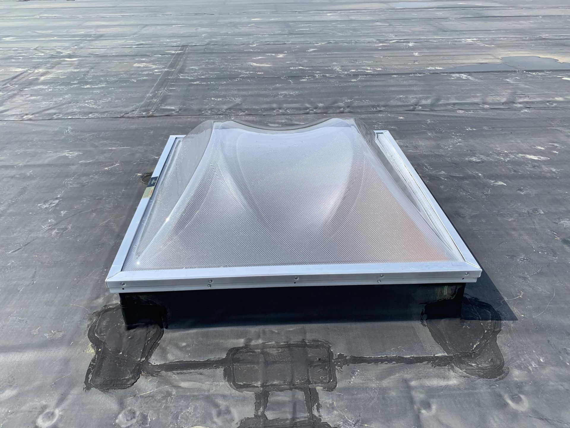 Skylight on a flat, black roof. Clear, textured dome. Silver frame around the base.