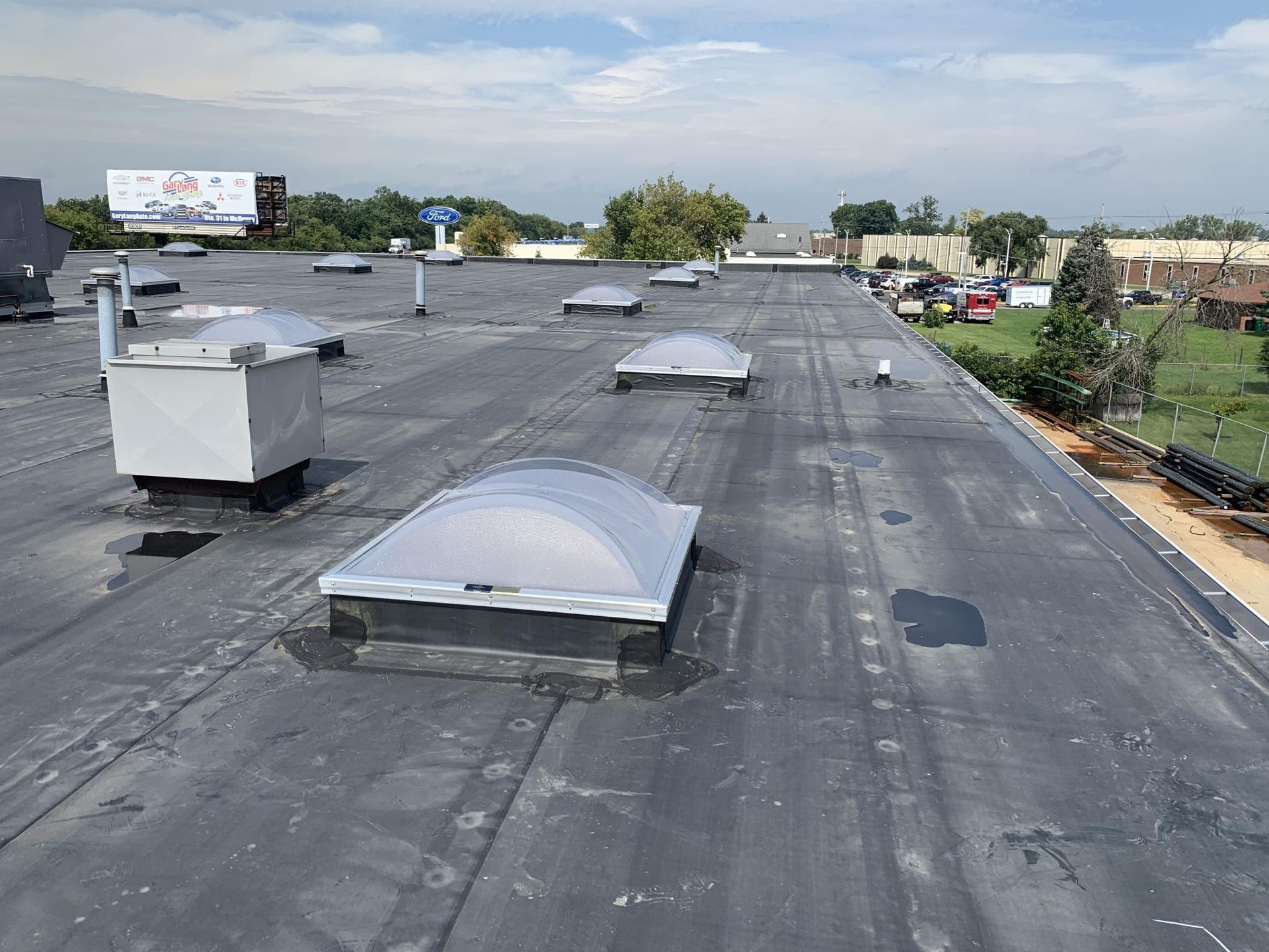A flat commercial roof with several skylights, vents, and a billboard in the distance on a sunny day.
