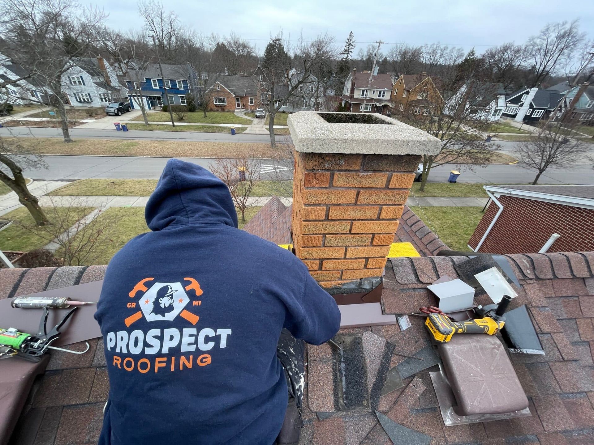 Roofer on a roof wearing a Prospect Roofing hoodie, working near a brick chimney on a cloudy day.