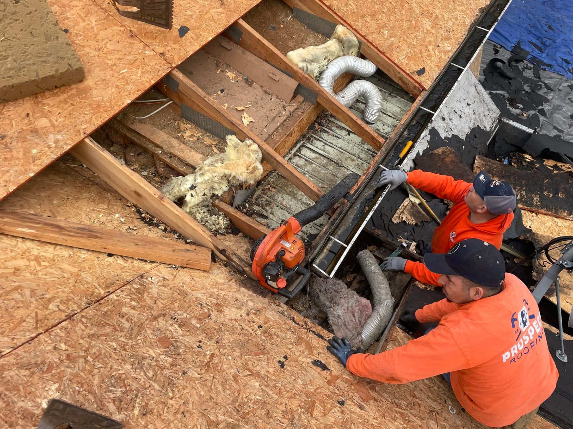 Two workers remove insulation from a roof opening, wearing orange shirts and black caps.