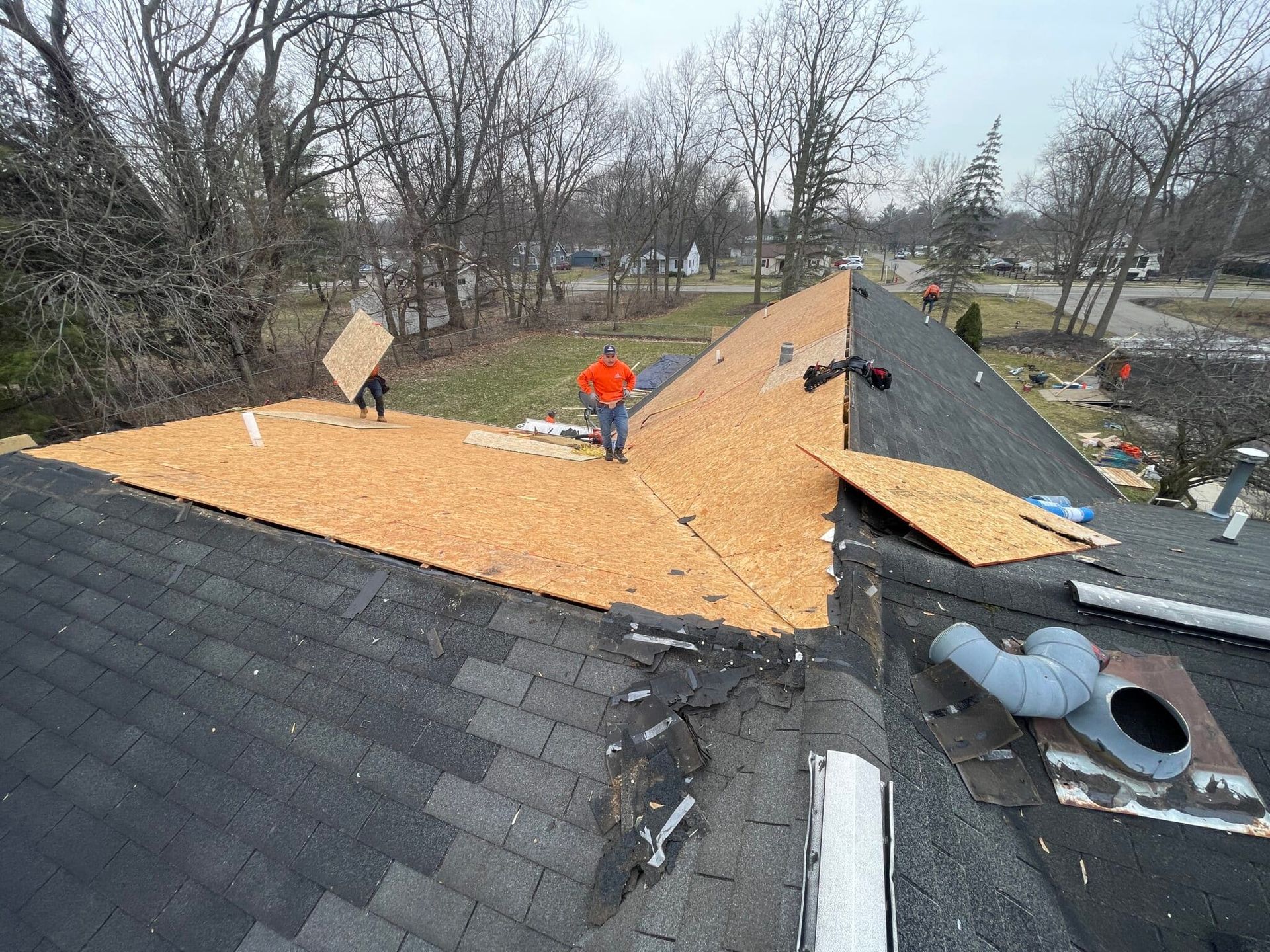 Workers replacing roof shingles on a house. New plywood on part of roof, old shingles on others. Overcast day.