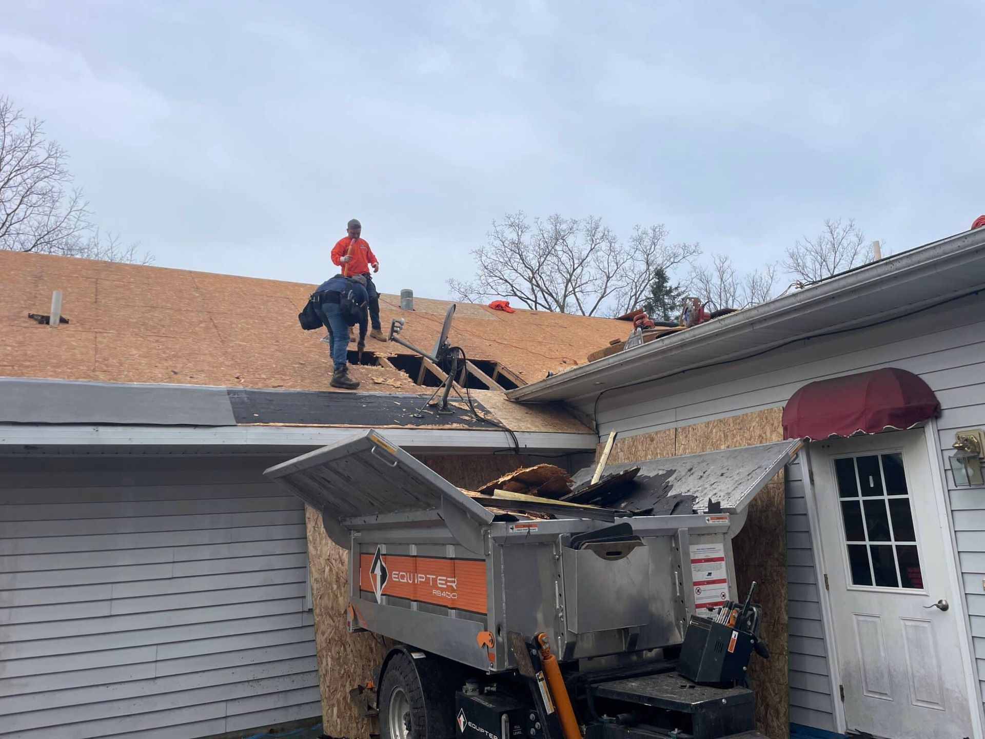 Roofers on a house partially missing a roof, debris in a dump truck, cloudy sky.
