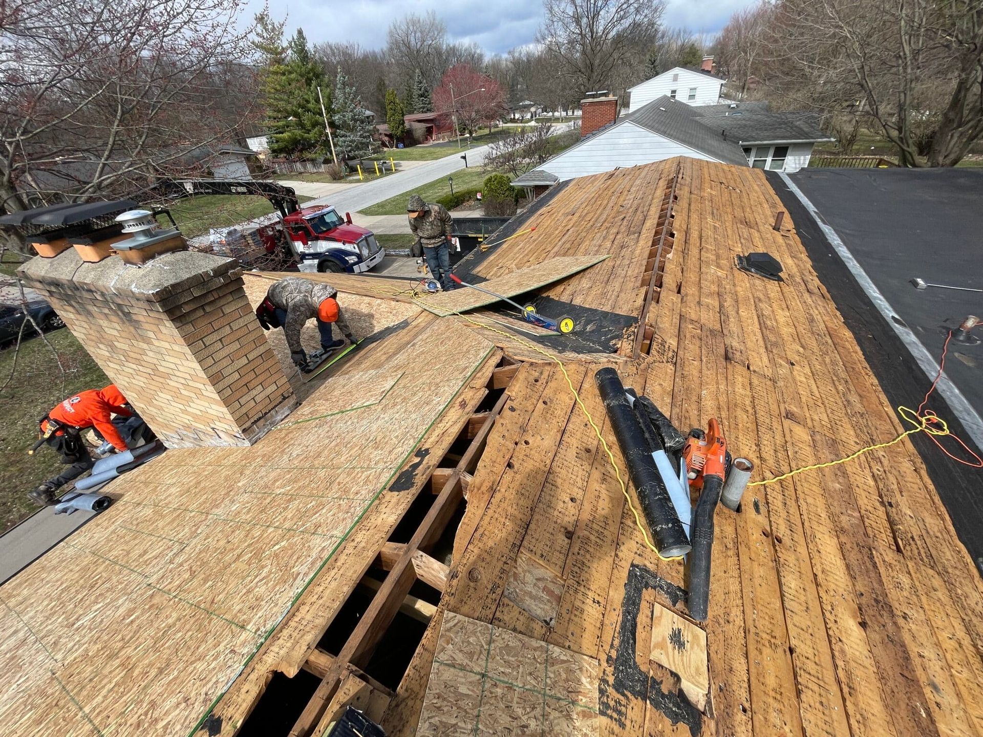 Roofers working on a partially shingled rooftop with wood panels, chimney, and surrounding neighborhood on a cloudy day.