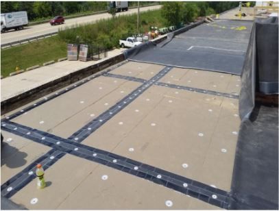 Rooftop construction: workers installing dark strips over beige material, next to a highway.