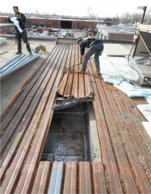 Two construction workers on a rooftop tearing up rusted metal roofing. Sunlight, city background.