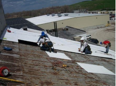 Workers installing white roofing material on a building with other buildings visible in the background.