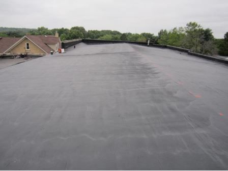 Black flat commercial roof, with a building in the background. Overcast sky and trees.