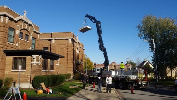 Crane lifting a rectangular object near a brick building; workers and traffic cones present.