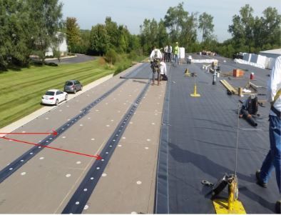 Workers on a flat roof installing dark roofing material with white circular fasteners, in a daylight setting.