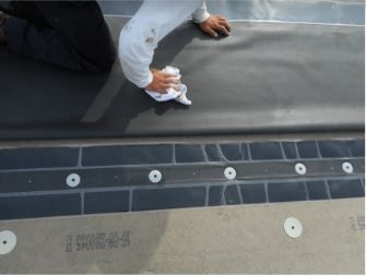 Person wiping sealant on a dark roofing membrane. Fasteners and joint details visible.