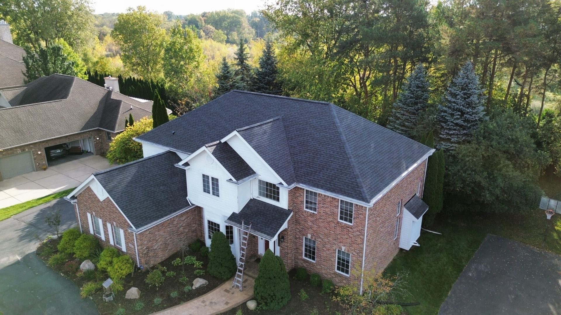 Two-story brick house with dark gray roof, surrounded by trees and landscaping, on a sunny day.