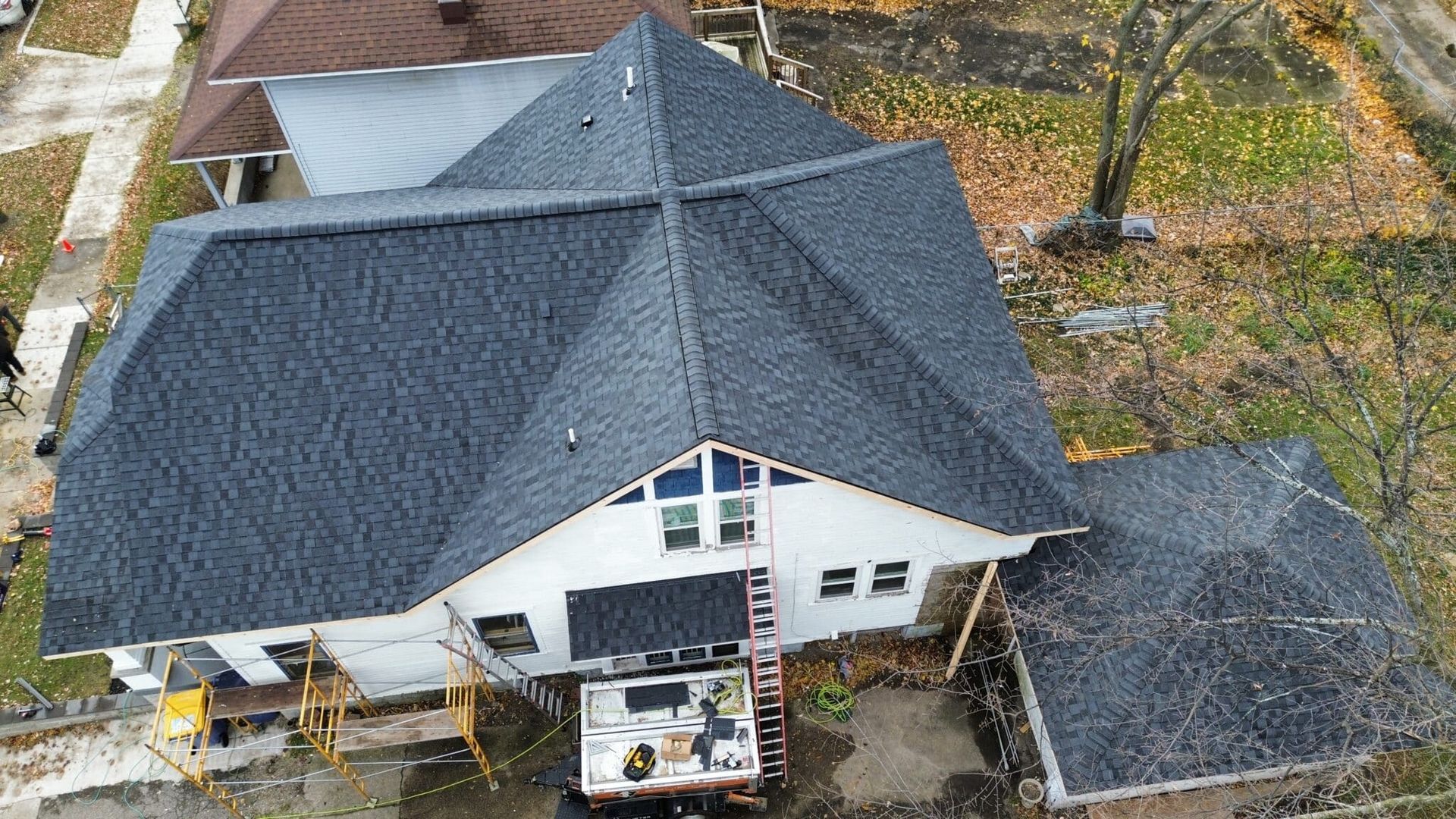 Aerial view of a house with a newly shingled dark roof, scaffolding, and a detached garage on a grassy lot.