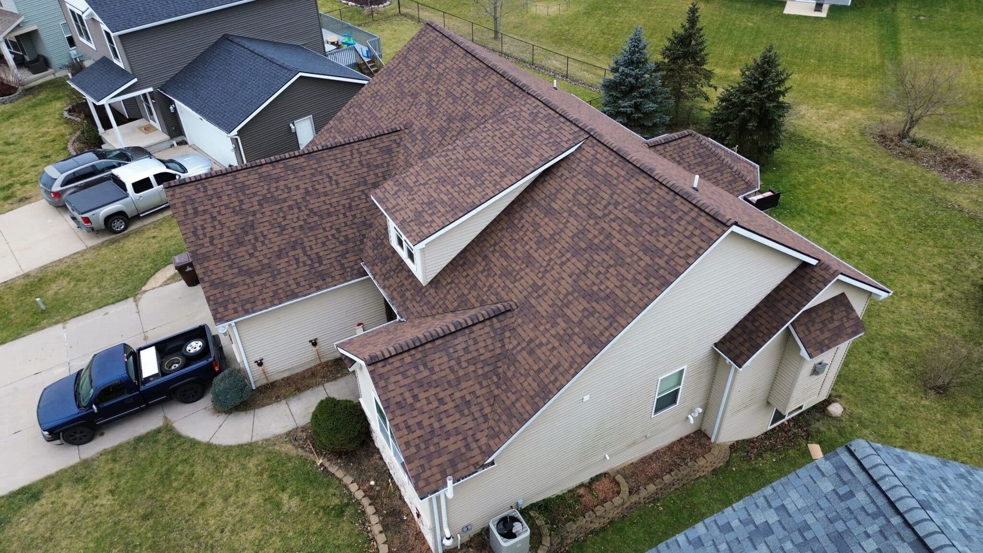Aerial view of a brown-roofed house with beige siding and two dormers. A blue truck is in the driveway.