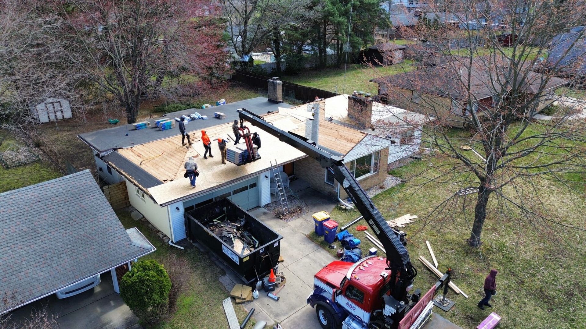 Workers replacing a roof on a house, with a crane lifting materials. A dumpster is on the driveway.