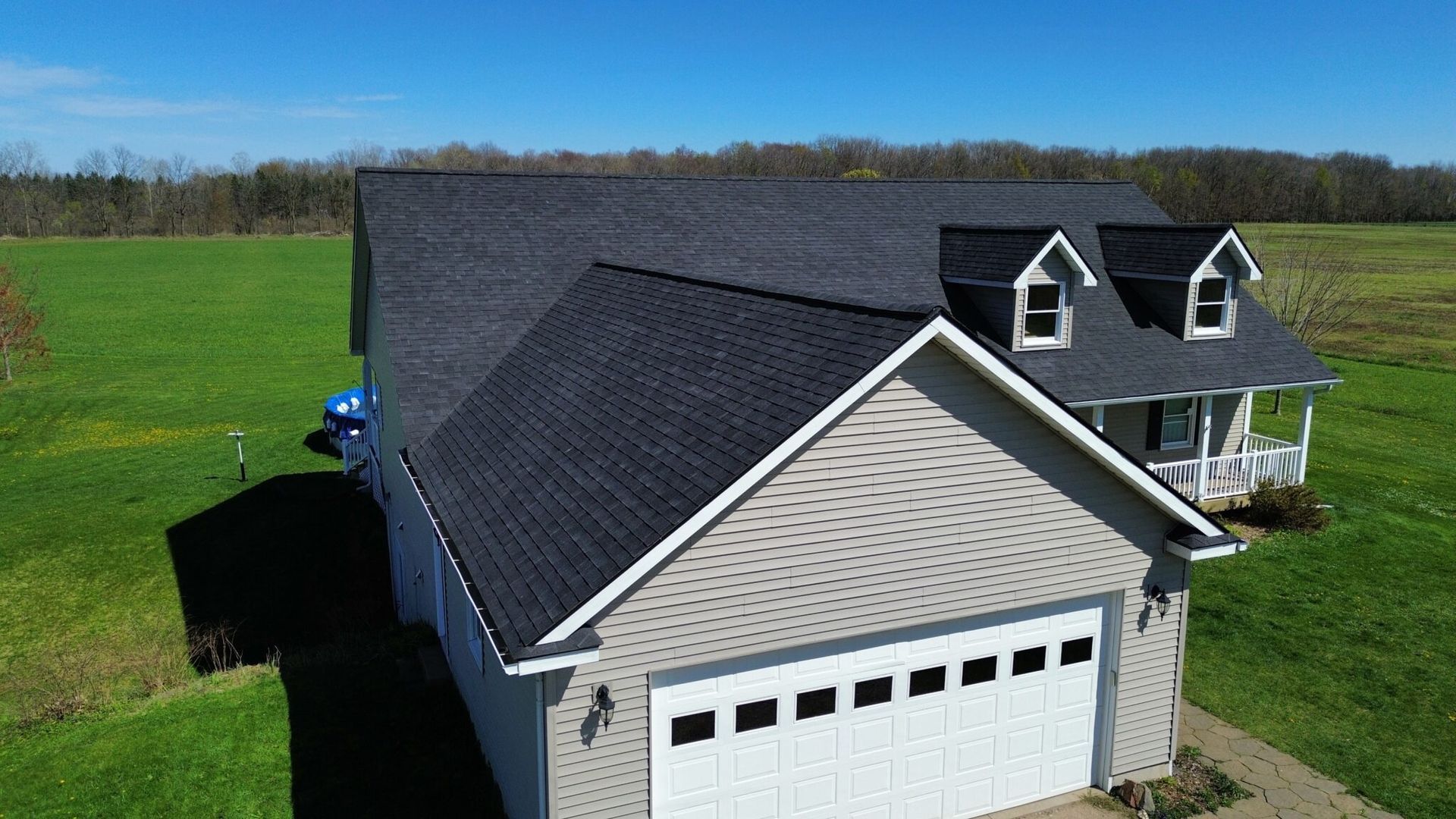 A house with a black roof, beige siding, and a white garage door, set in a green field.