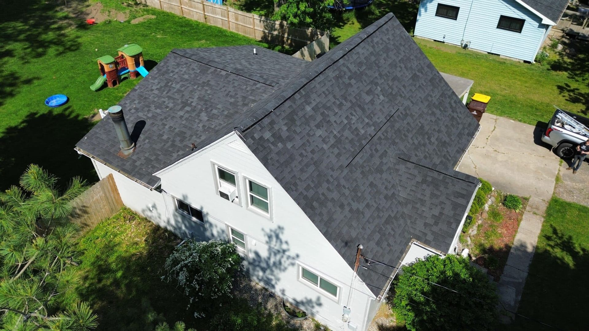 Overhead view of a two-story house with dark gray roof, surrounded by trees and grass; blue and white house in the background.
