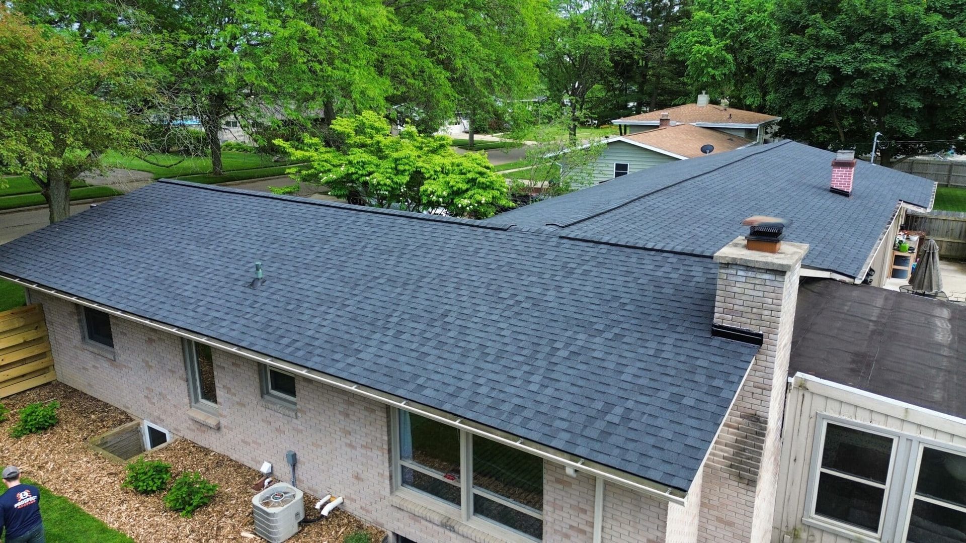 Dark blue asphalt shingle roof on a brick house; green trees in the background.