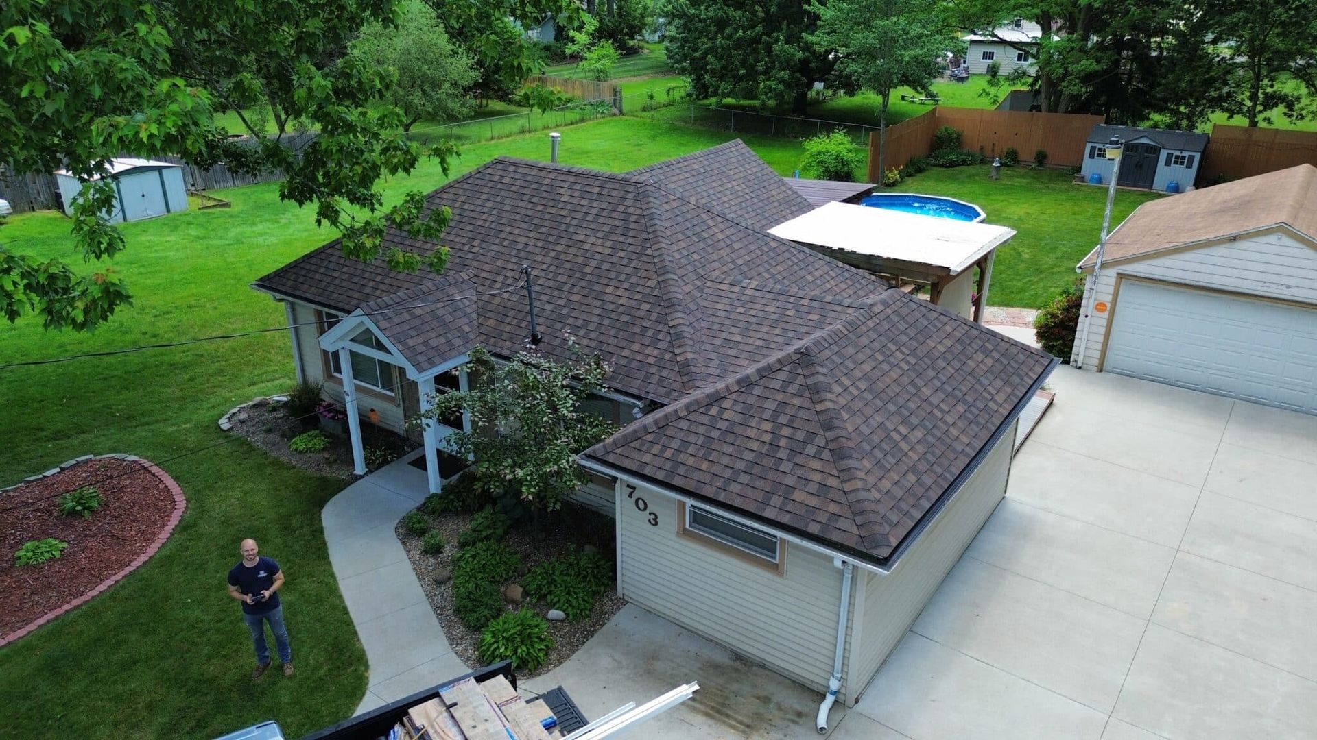 Aerial view of a house with a brown shingle roof, green lawn, and a person standing outside holding a drone controller.