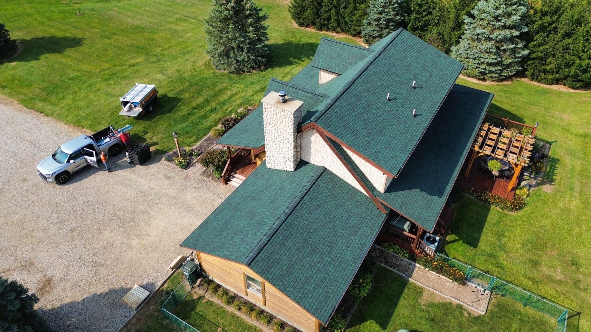 Aerial view of a house with green roof, chimney, and truck parked on a gravel driveway, in a grassy setting.
