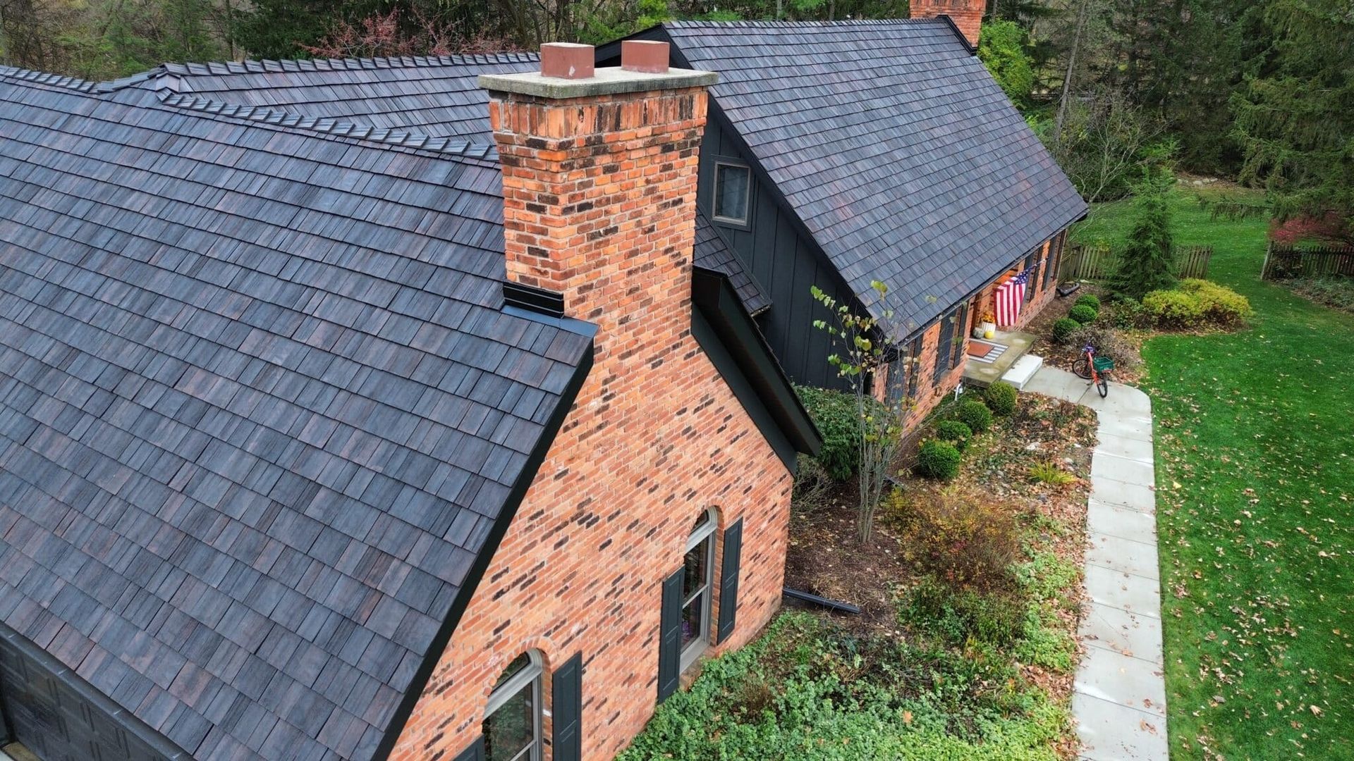 Dark shingle roof on a house with a brick chimney. Green lawn and a concrete path.