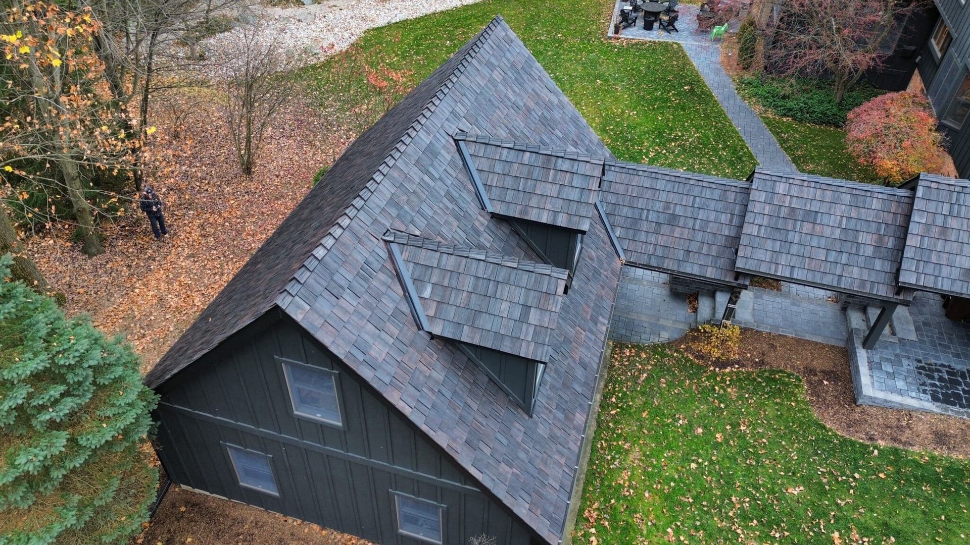 Overhead view of a dark-roofed building with a walkway, surrounded by trees and grass covered in autumn leaves.