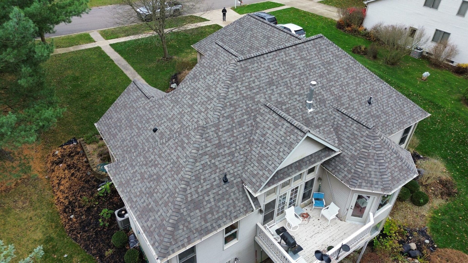 Aerial view of a gray-roofed house with a deck, surrounded by a grassy yard and trees.