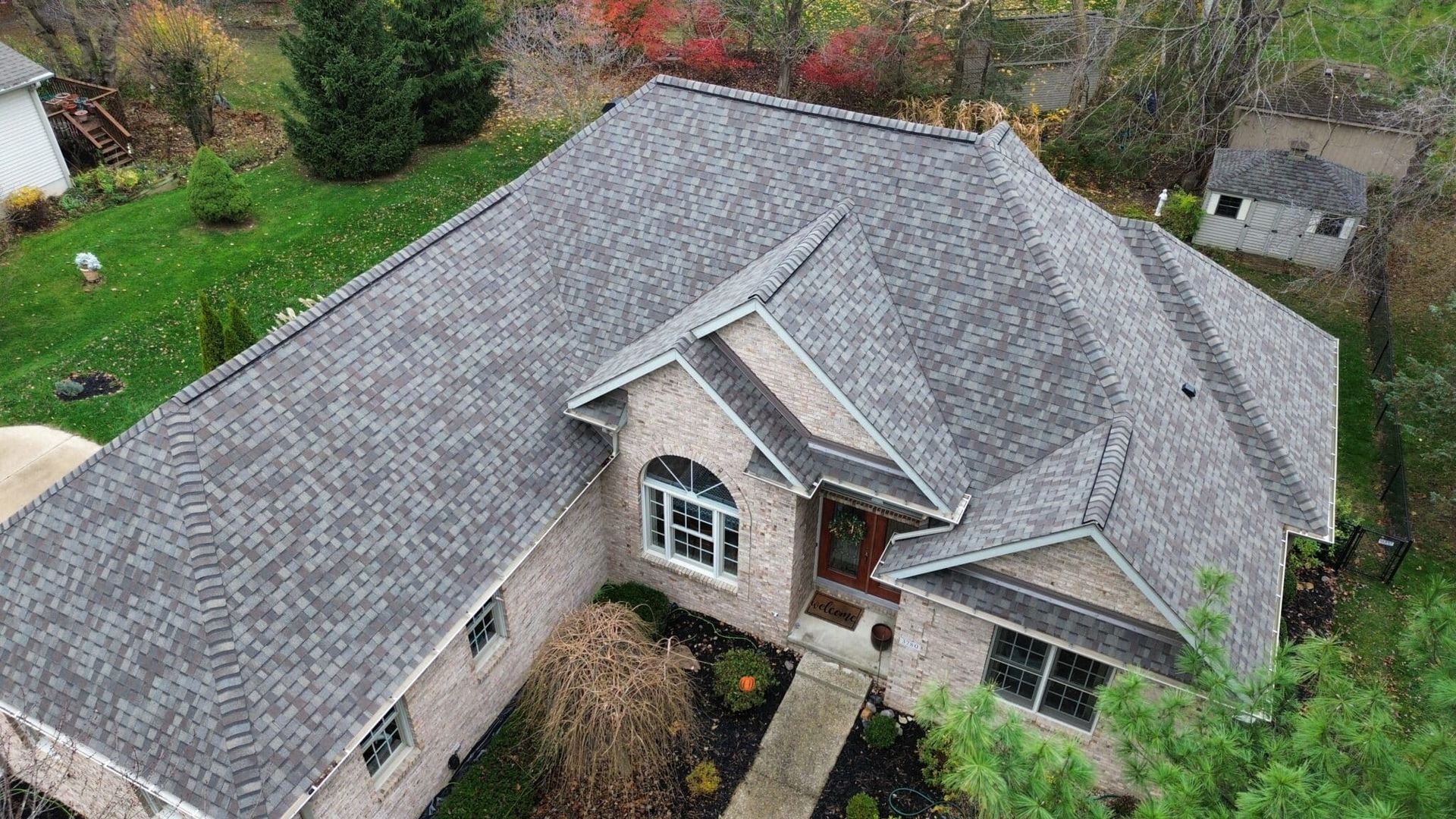 Aerial view of a gray-shingled roof on a brick house with green grass and trees in the background.