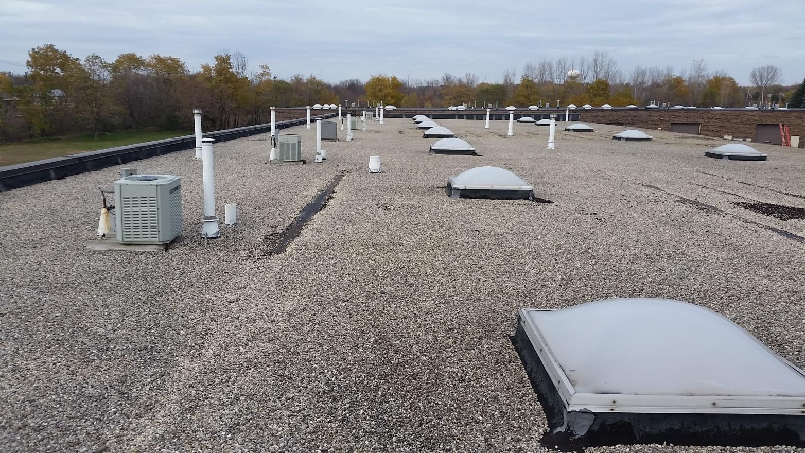 Gravel-covered flat commercial roof with HVAC units, skylights, and pipes against a cloudy sky.